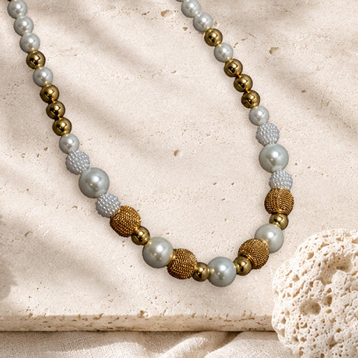 A beaded necklace with white and creme beads, textured gold beads, and metallic gold beads, styled on a beige stone slab with seashells, coral, and dried palm leaves.