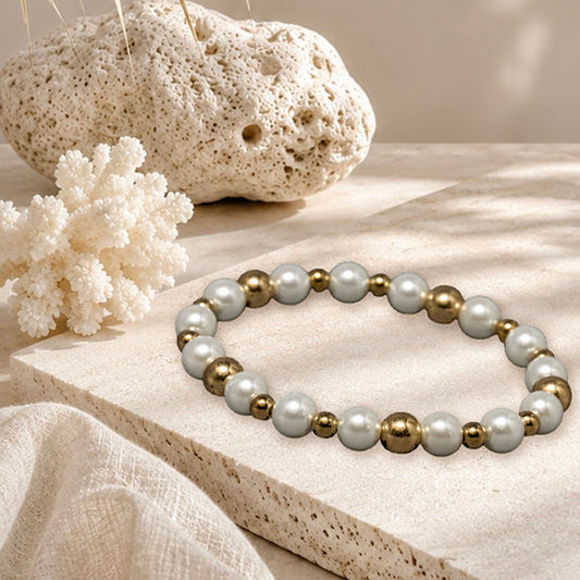 Beaded stretch bracelet with white pearls and metallic gold spacer beads, displayed on a textured stone slab with beach-themed props like a seashell, coral, and palm leaves.