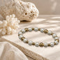 Beaded stretch bracelet with white pearls and metallic gold spacer beads, displayed on a textured stone slab with beach-themed props like a seashell, coral, and palm leaves.