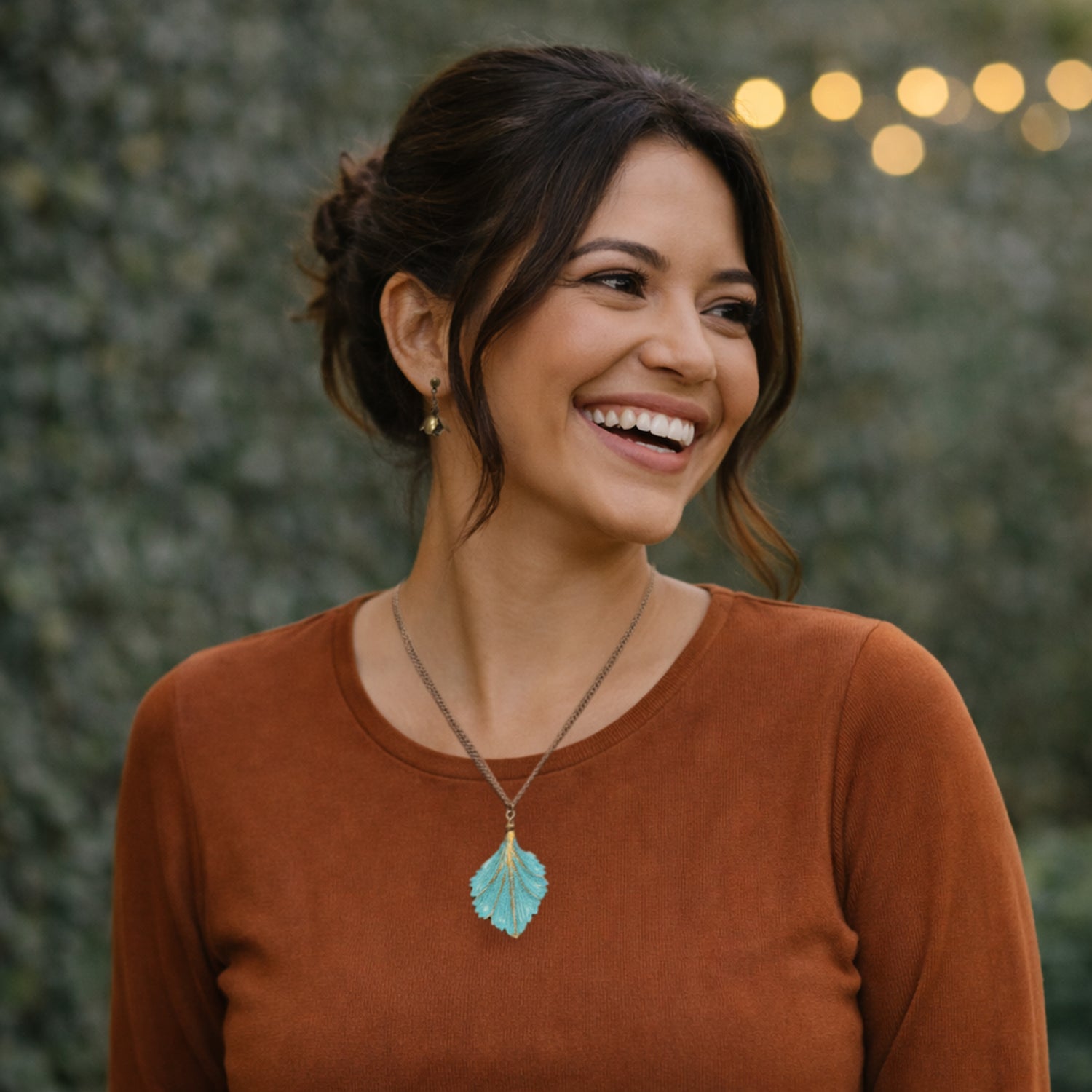 Woman wearing a brown shirt with a necklace featuring a petnia verdigris leaf pendant, standing against a blurred green background.