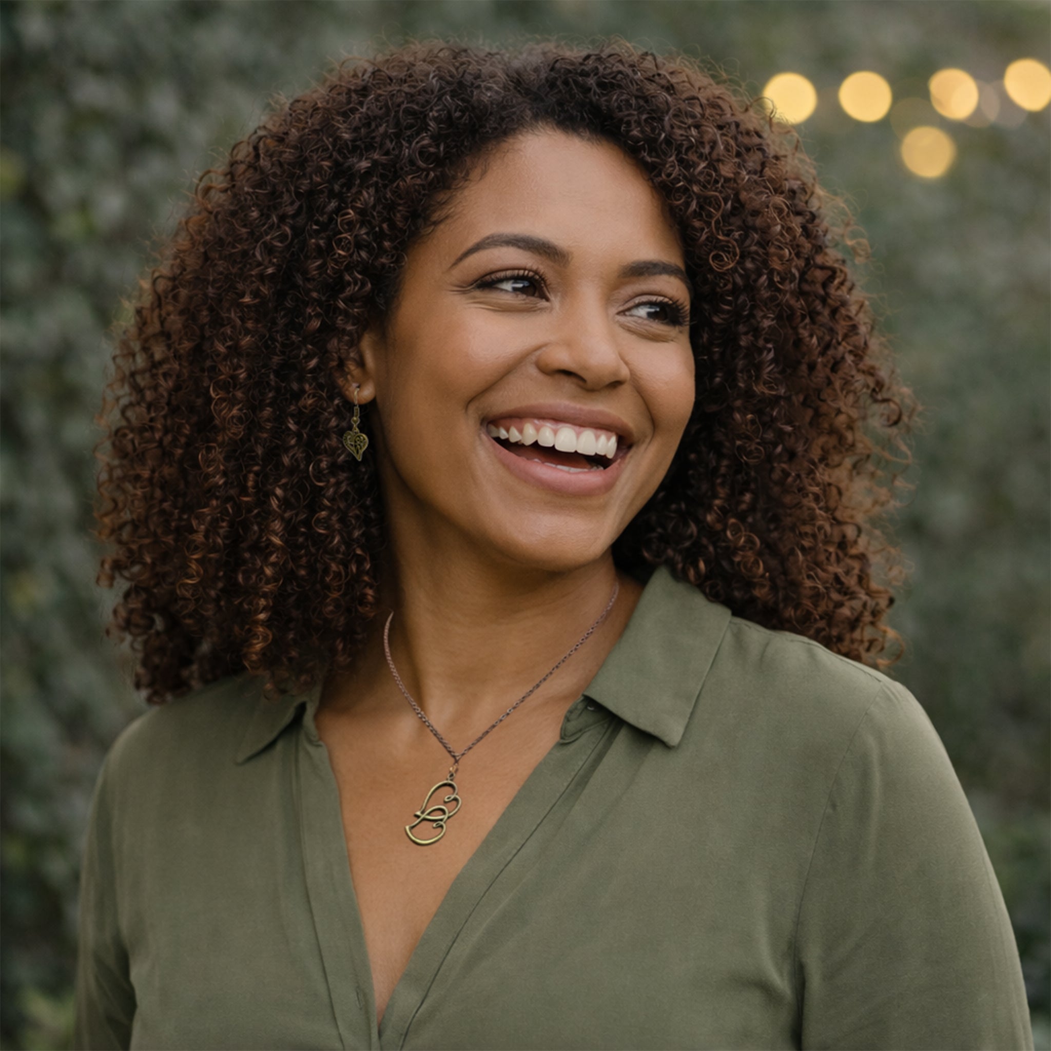 Woman with curly hair wearing a green shirt and necklace, standing outdoors with blurred lights in the background.