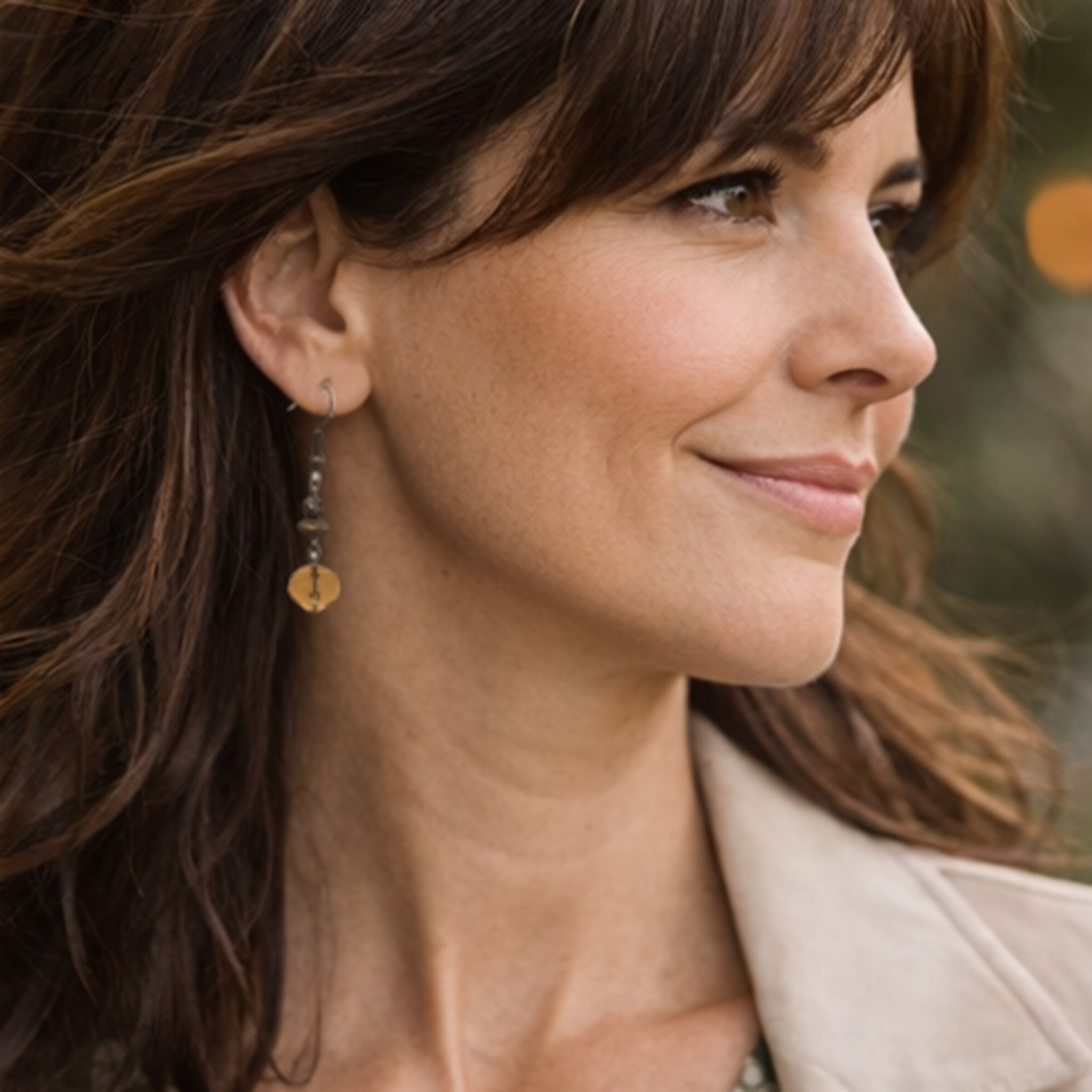 Woman with long brown hair wearing earrings, with a blurred natural background
